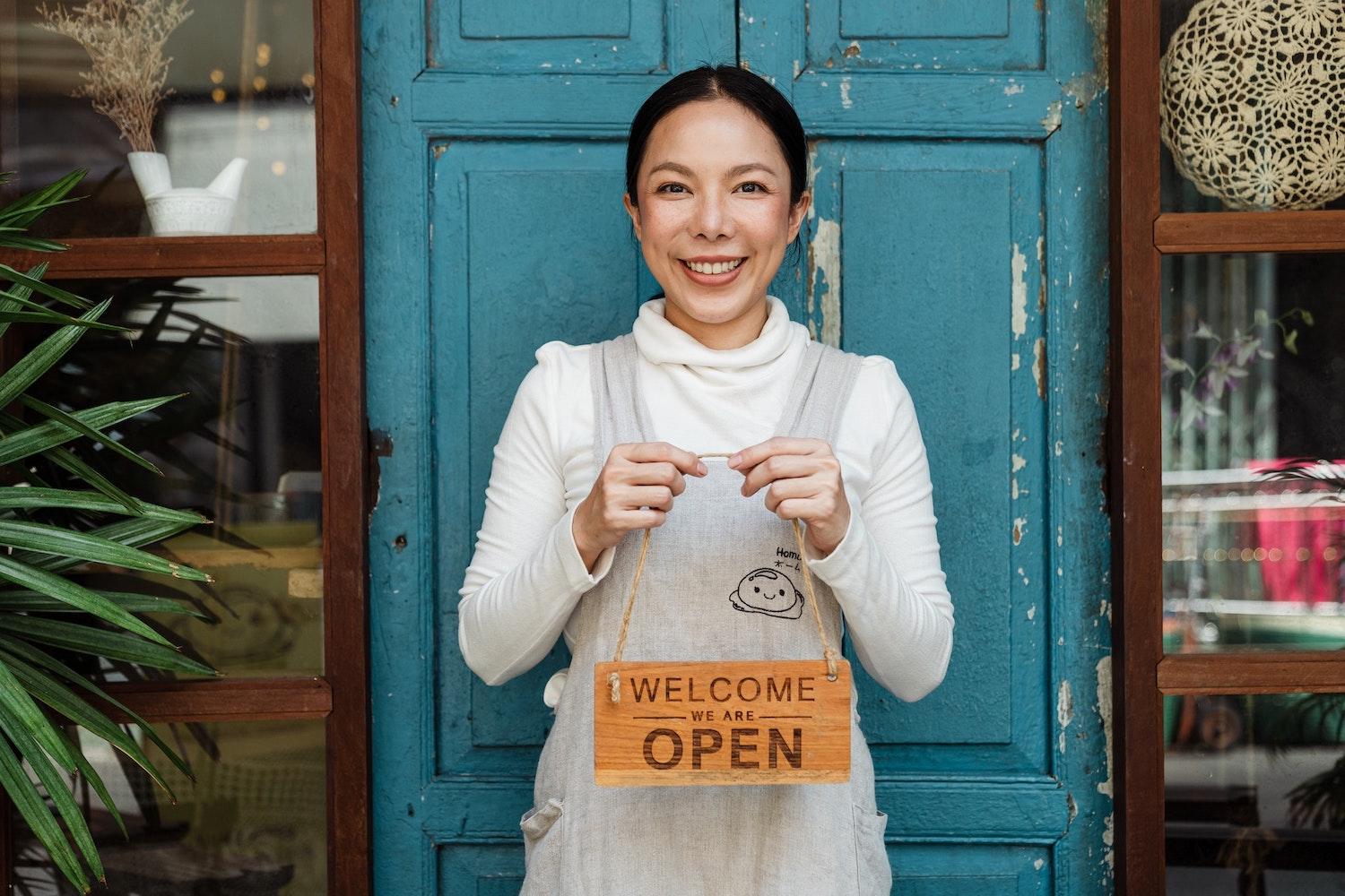 Image of woman standing in front of small business with a sign reading 'welcome we are open'