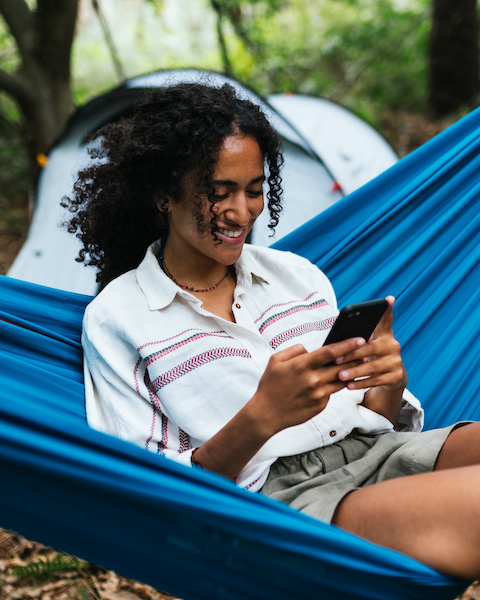 Cheerful curly hair woman smiling and laughing while she is using her smartphone, seated on a blue hammock. Camping concept.